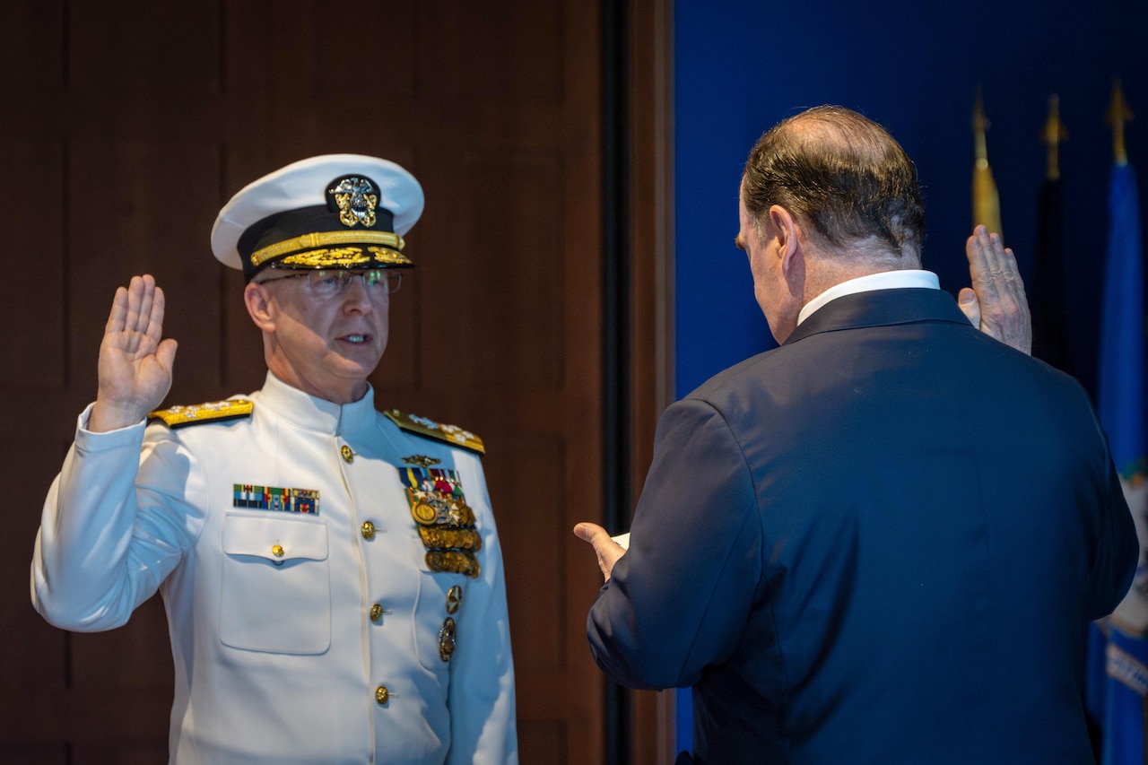 A man wearing a formal military uniform raises his right hand and speaks in front of a man in business attire, also with his right hand raised.