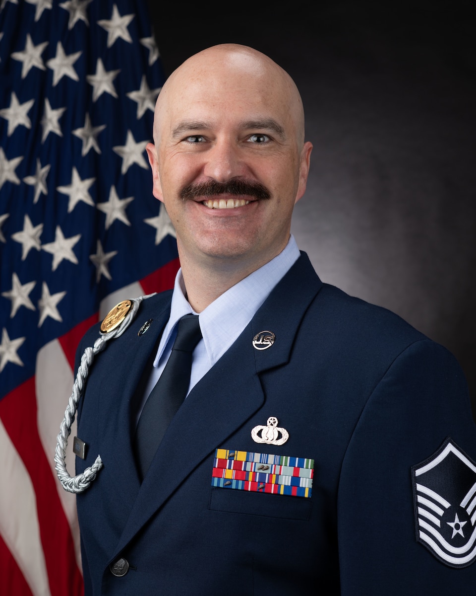 Official Photo of MSgt Benjamin Huseby, trombone with the USAF Bands.  MSgt Huseby is wearing blue service dress in front of the American flag.