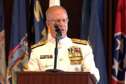 A man wearing a formal military uniform speaks into a microphone from behind a podium with flags behind him.