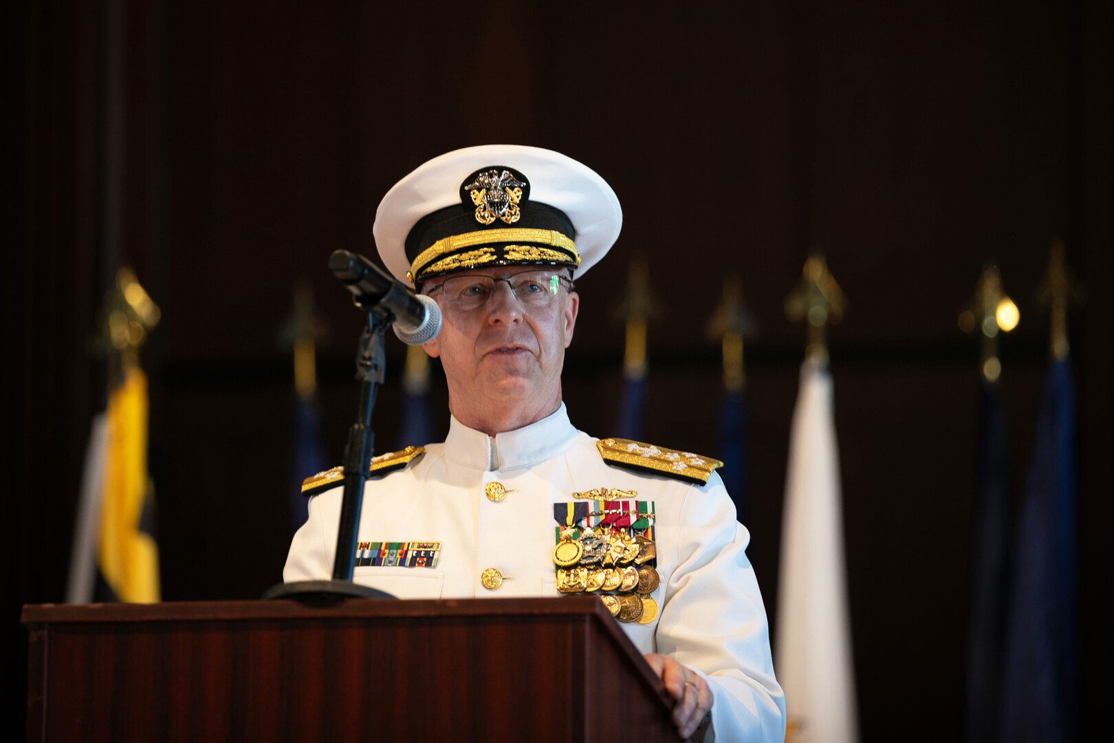 A man wearing a formal military uniform speaks into a microphone from behind a podium with flags behind him.