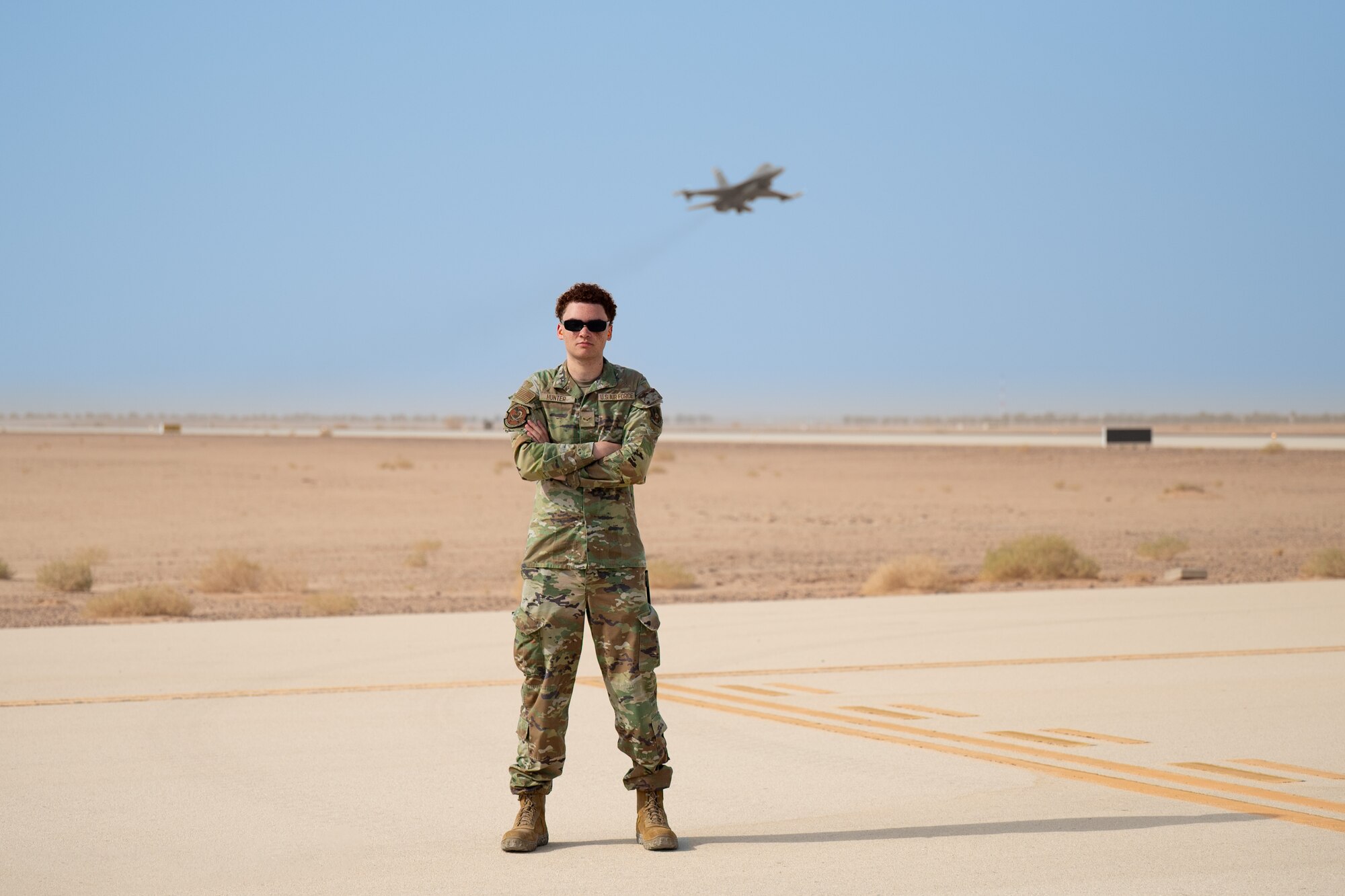 Airman poses on the flightline as an F-16C takes off behind him