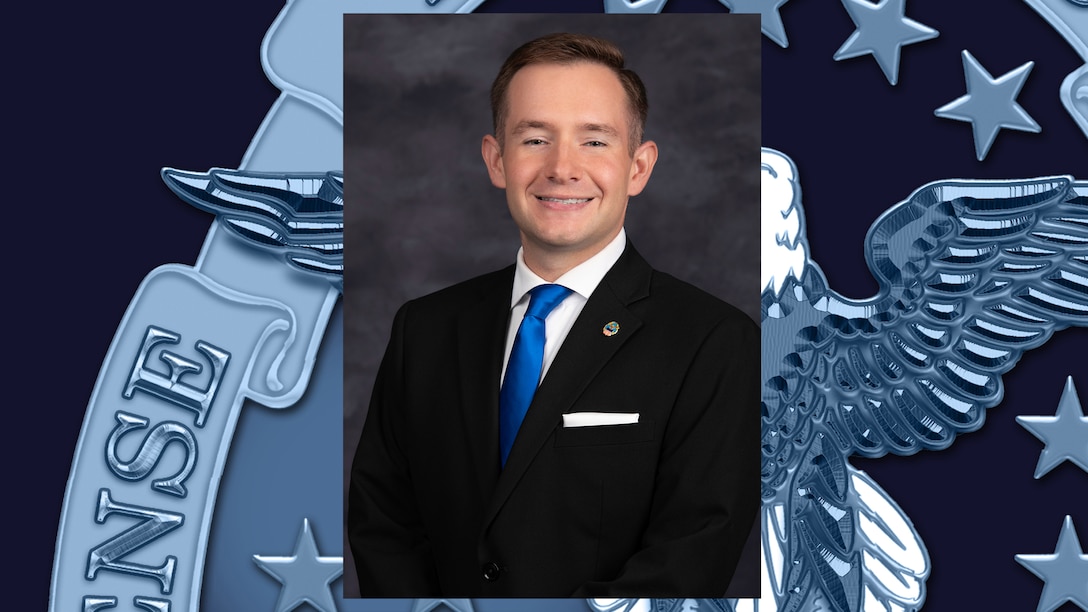 headshot of man in black suit, white shirt and blue tie