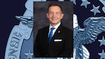 headshot of man in black suit, white shirt and blue tie