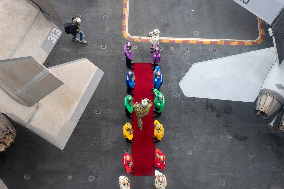 Sailors in colored shirts stand and salute, flanking a red carpet where a sailor walks on a ship's deck shown from above.