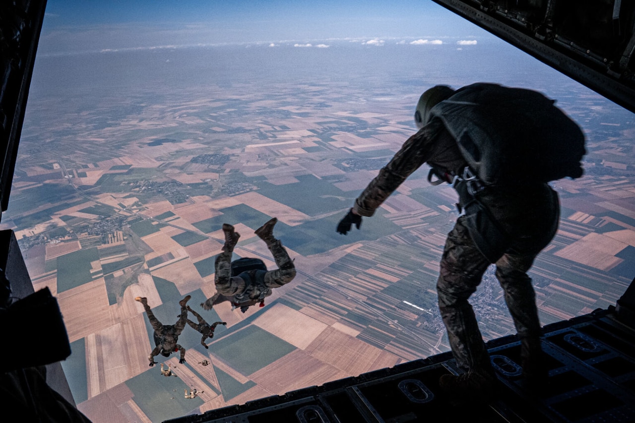 Several people in camouflage military uniforms and parachutes jump out of a plane. The aerial view of a landscape full of fields and small towns is in the background.