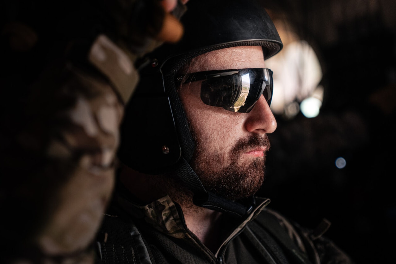 A bearded man wearing a helmet and sunglasses looks to the right.