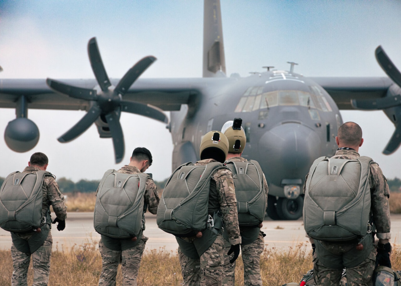 Several men in camouflage military uniforms and parachutes walk in a field with their backs to the camera. In the background is a military aircraft.