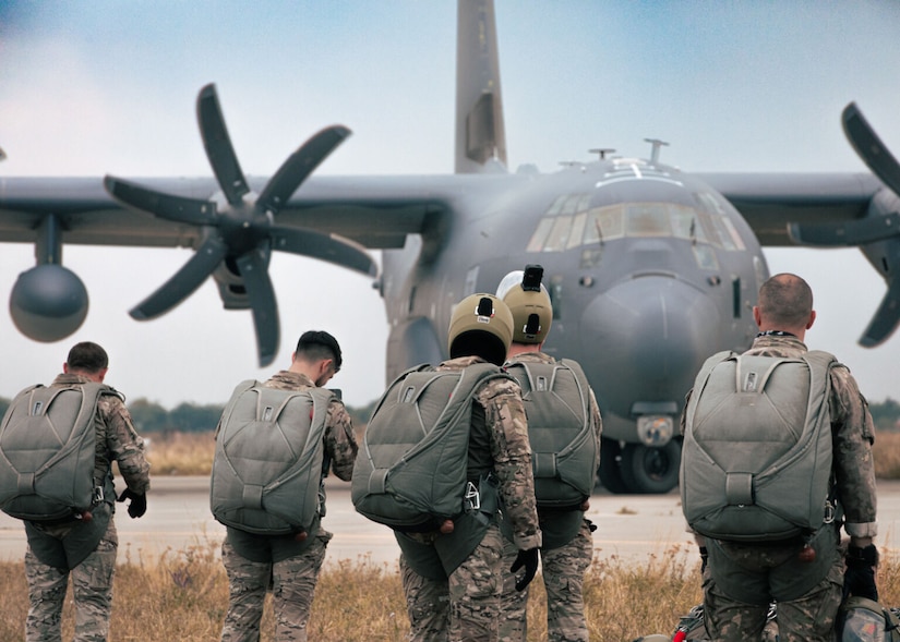 Several men in camouflage military uniforms and parachutes walk in a field with their backs to the camera. In the background is a military aircraft.