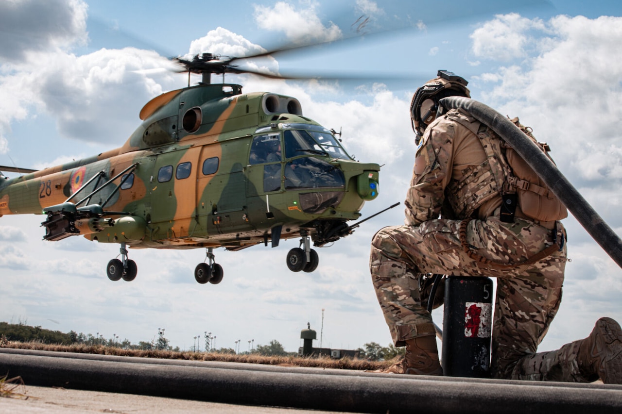A person in a camouflage military uniform kneels on the ground with a hose over their shoulder. A helicopter is hovering off the ground to the left of the person.