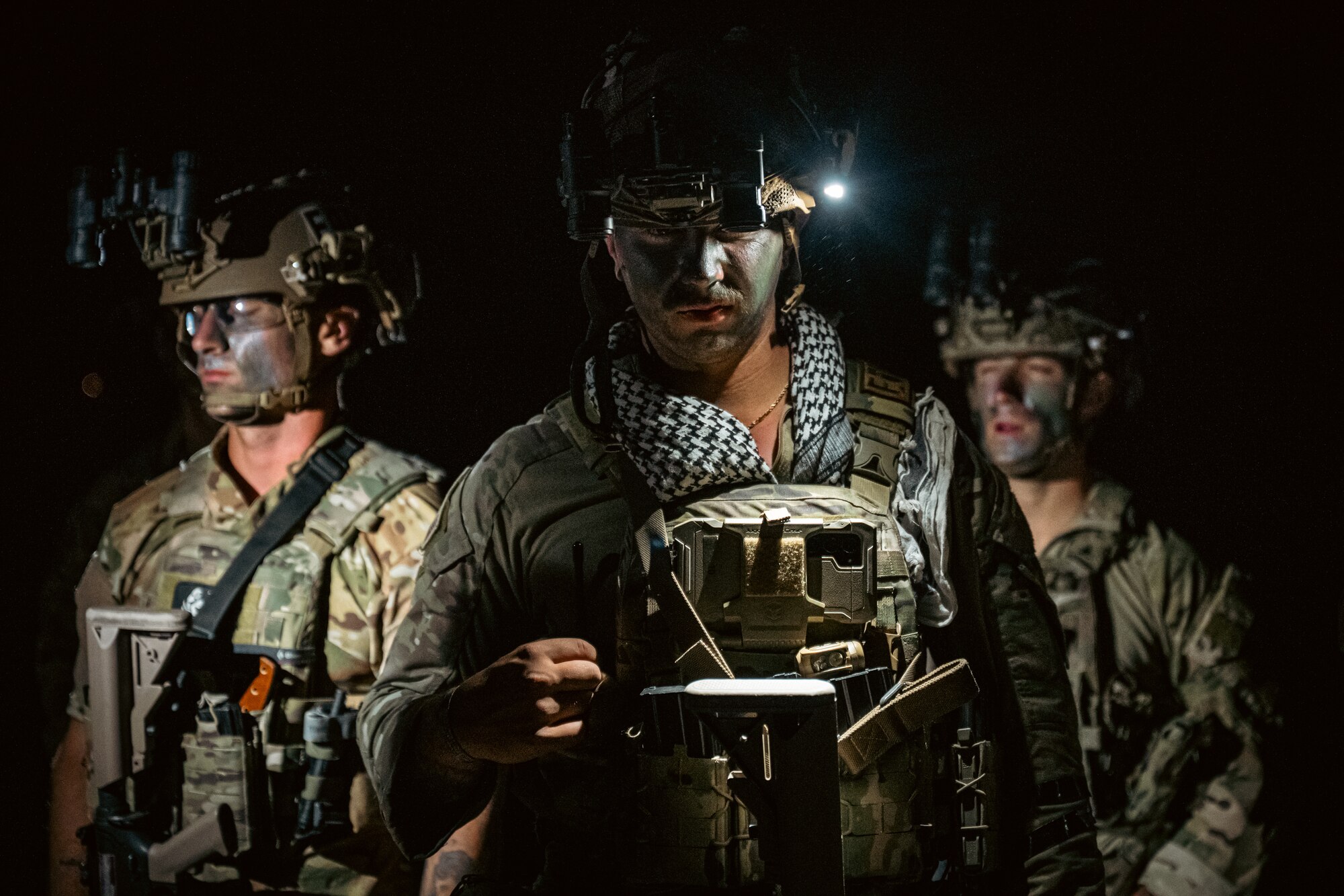 Three Airmen in combat gear in the dark with light shining on them.