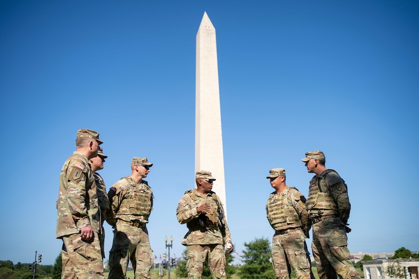 Men in camouflage military uniforms stand in a semicircle speaking to each other. The Washington Monument is in the background.