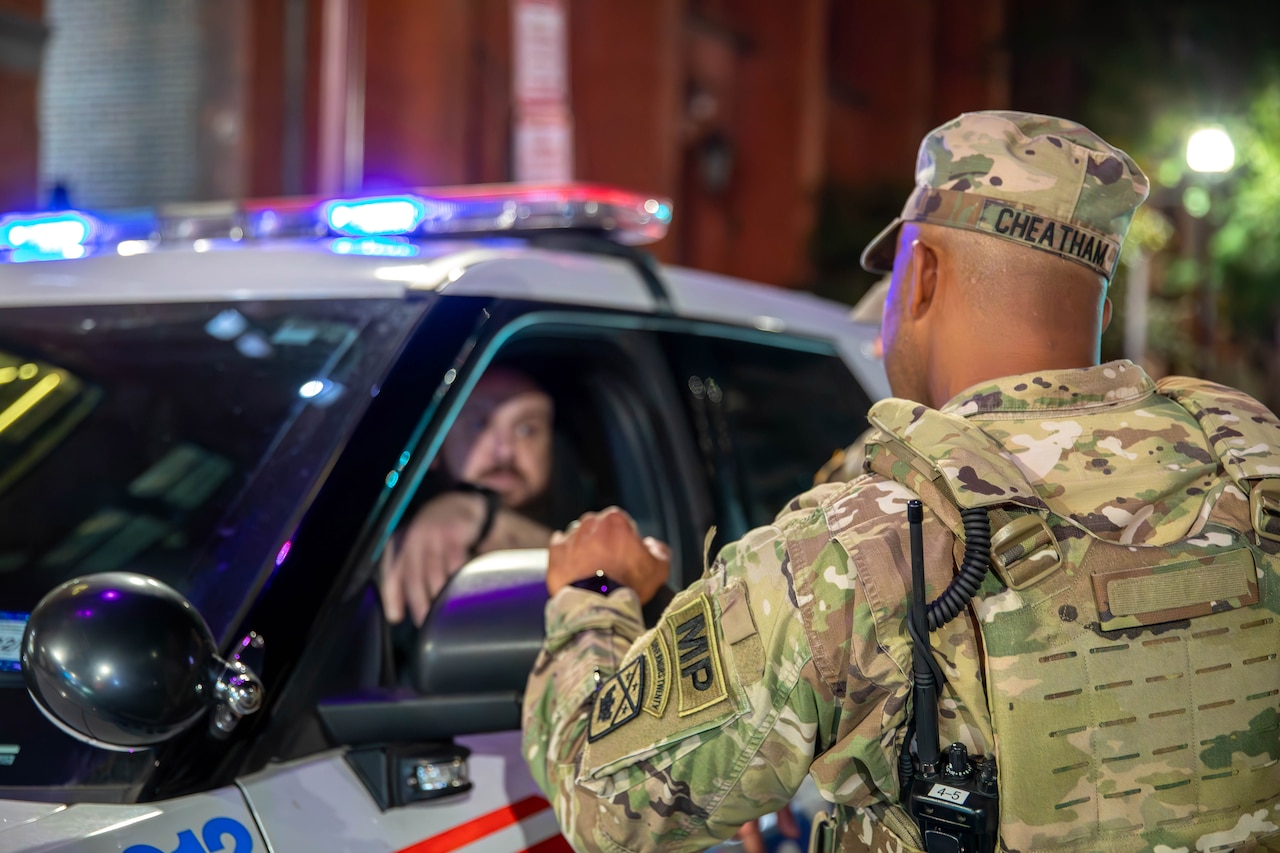 A man in a camouflage military uniform speaks to a man in a police vehicle.