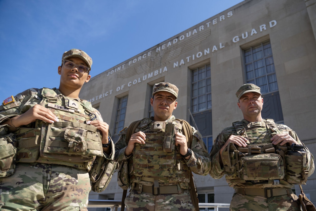 Three people in military camouflage uniforms stand together outside in front of a building. The top of the building says, Joint Force Headquarters, District of Columbia National Guard.