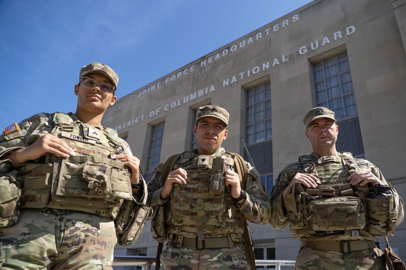 Three people in military camouflage uniforms stand together outside in front of a building. The top of the building says, Joint Force Headquarters, District of Columbia National Guard.