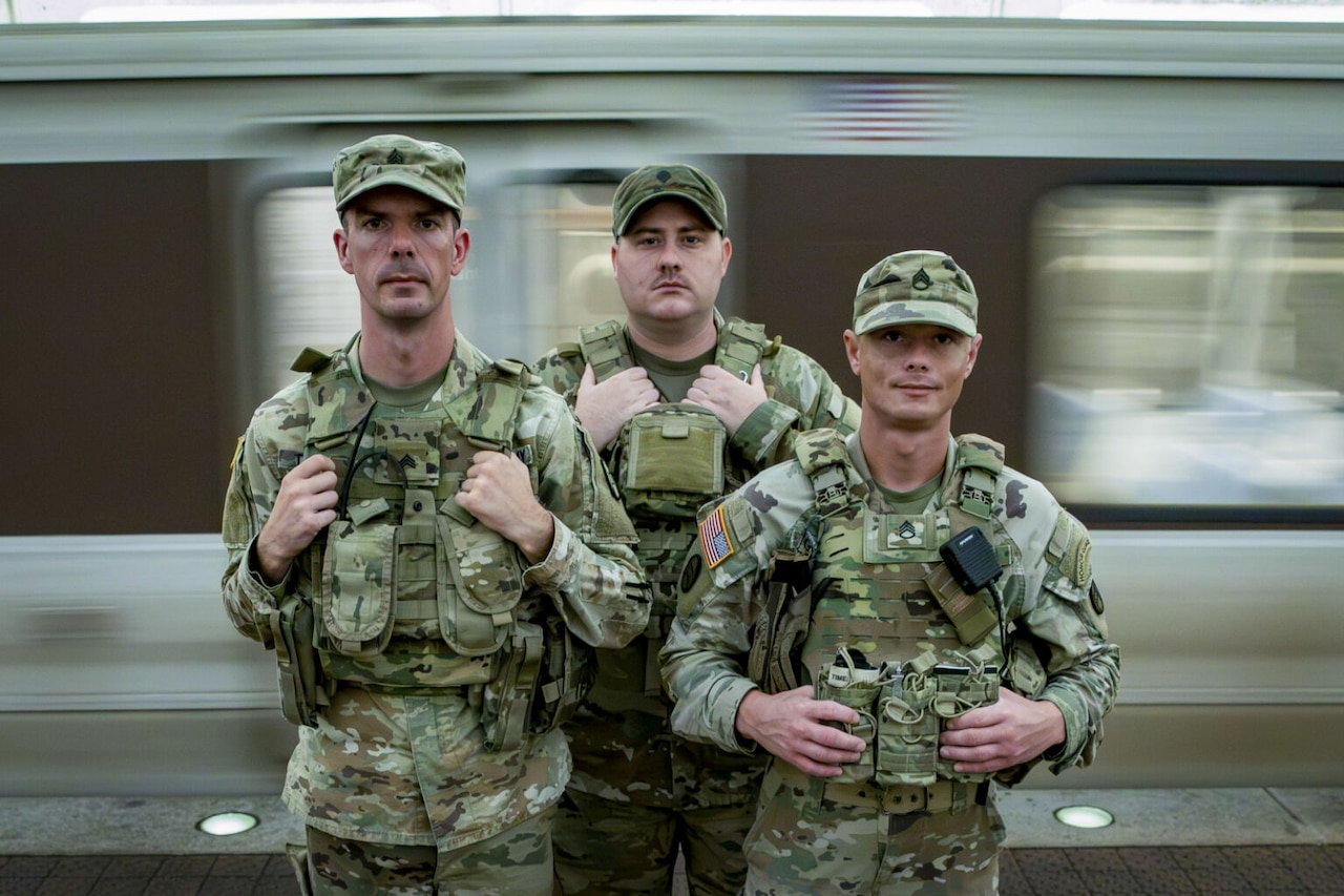 Three people in military camouflage uniforms stand together in an underground train station as a train moves behind them.