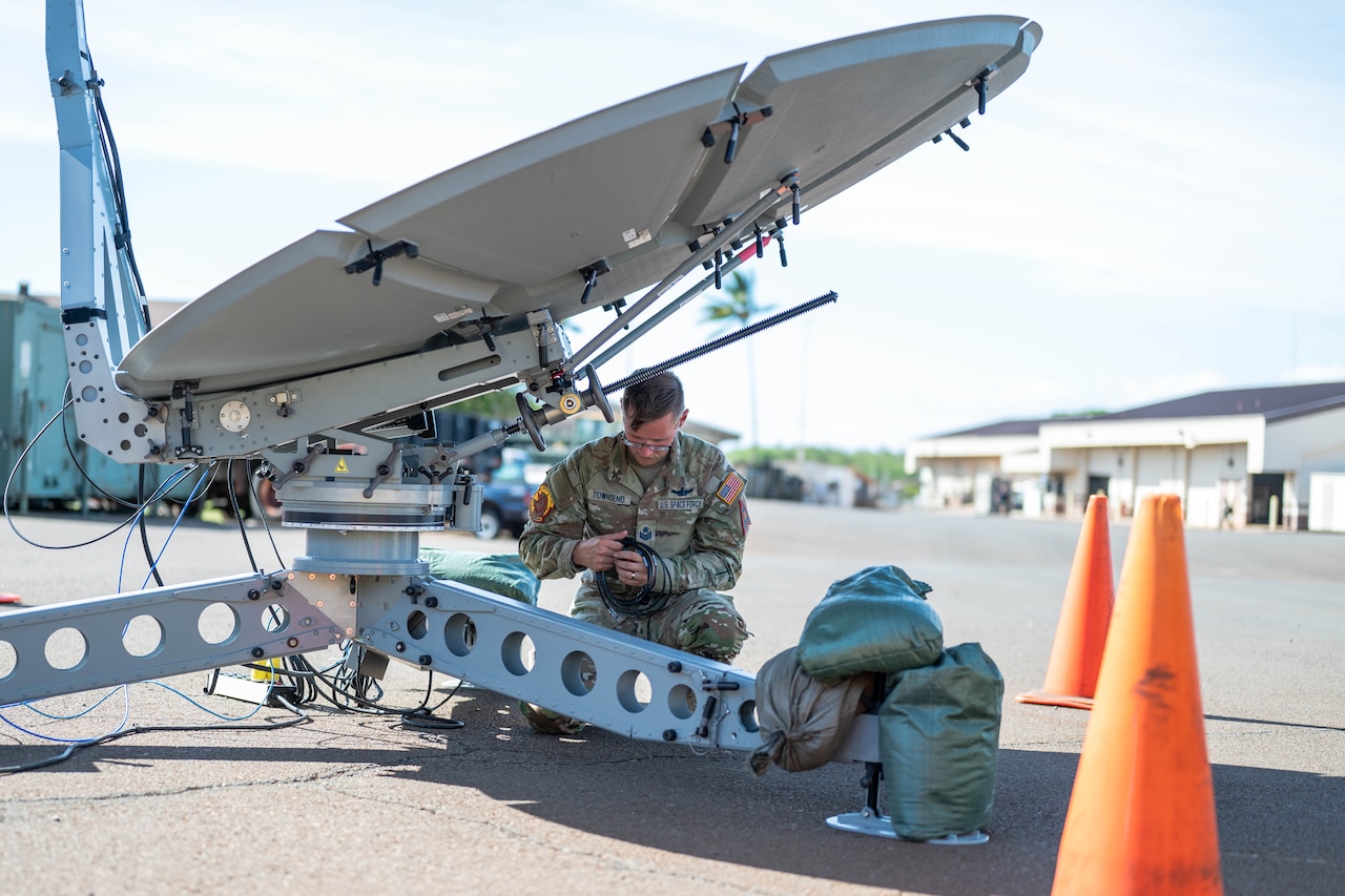 A person in a camouflage military uniform unrolls a cable while crouching under a large satellite dish. There are orange cones placed around the dish and sandbags on its platform.
