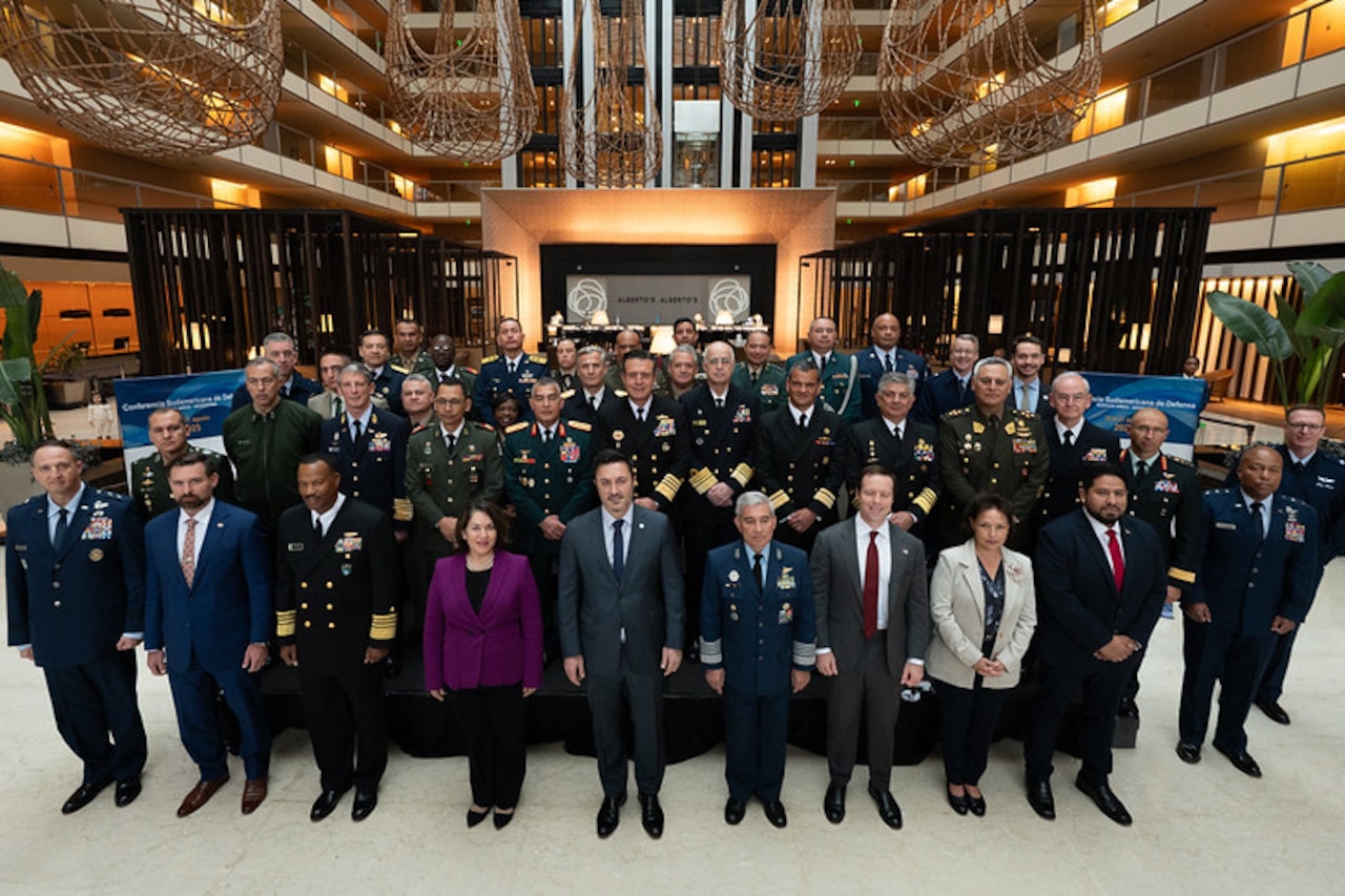 Dozens of people in military dress uniforms and business attire stand in rows while posing for a photo.