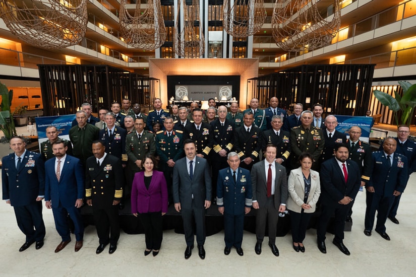 Dozens of people in military dress uniforms and business attire stand in rows while posing for a photo.