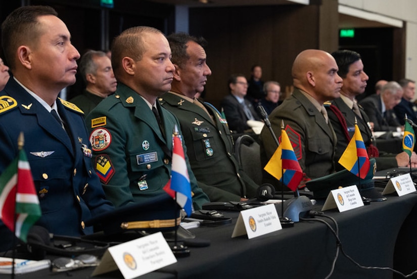 People in military dress uniforms sit behind a table with small national flags in front of them. There are several people in business attire seated in the background.