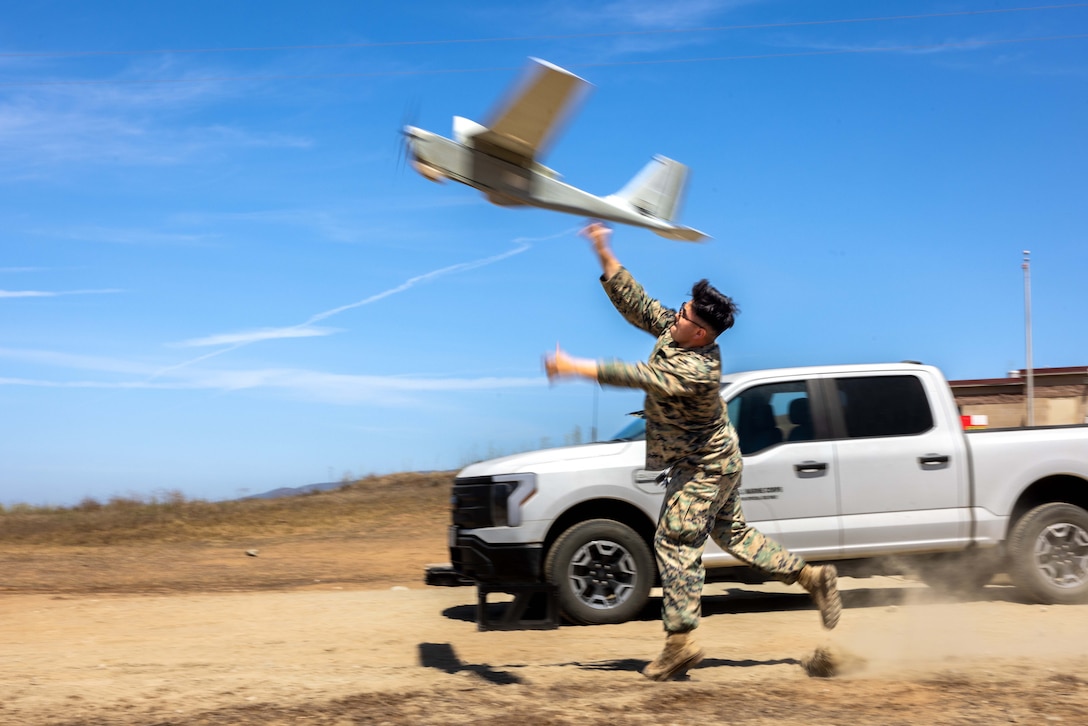 A Marine runs on a dirt path next to a white truck and releases a small unmanned aircraft system into a blue sky.