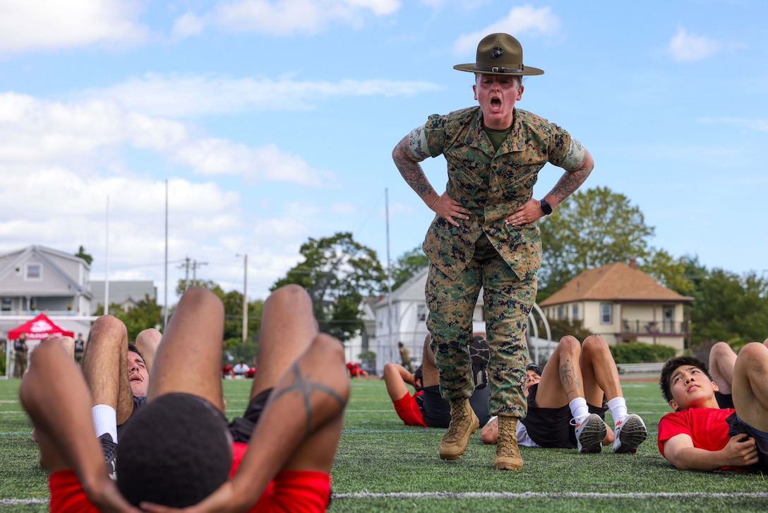 A Marine stands and yells down at poolees who are wearing red shirts and doing crunches on a field during the day