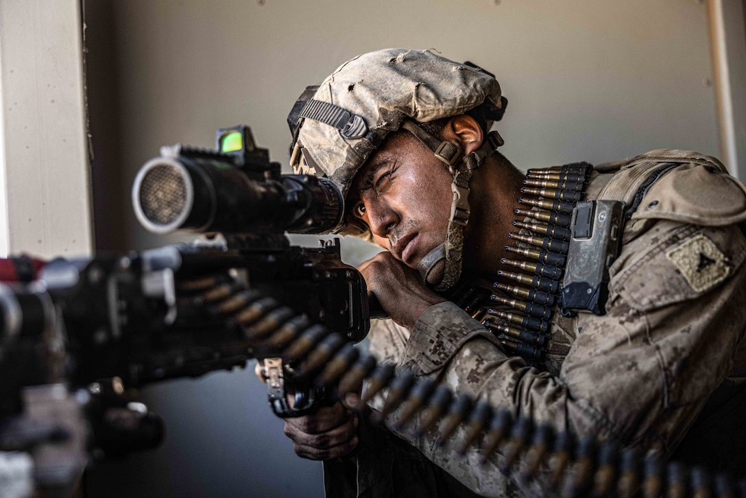 A Marine in tactical gear with a bullet belt draped over their shoulder looks through the scope of a service weapon in a room.