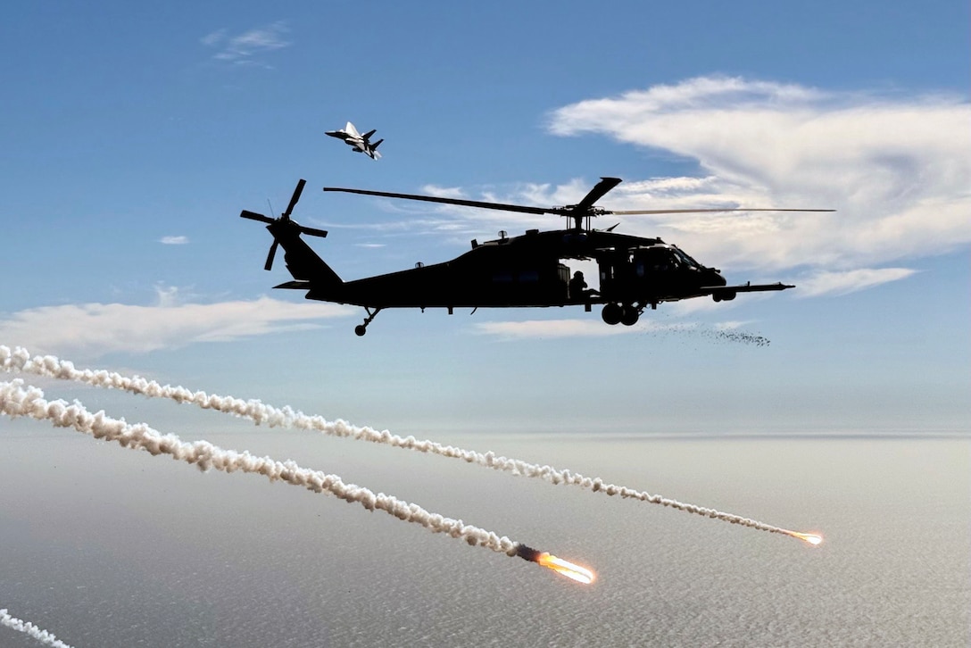 A helicopter flies in a blue sky above a body of water as an aircraft flies in the background and flares fly to the right.
