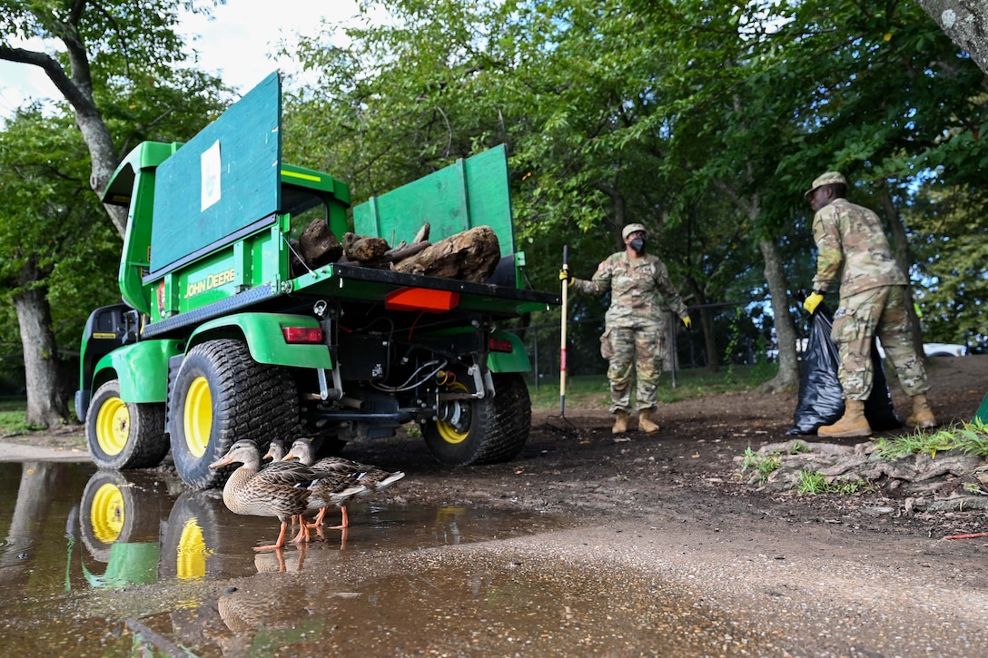 Two guardsmen collect trash next to trees and a green vehicle while three ducks stand in a puddle to the left.