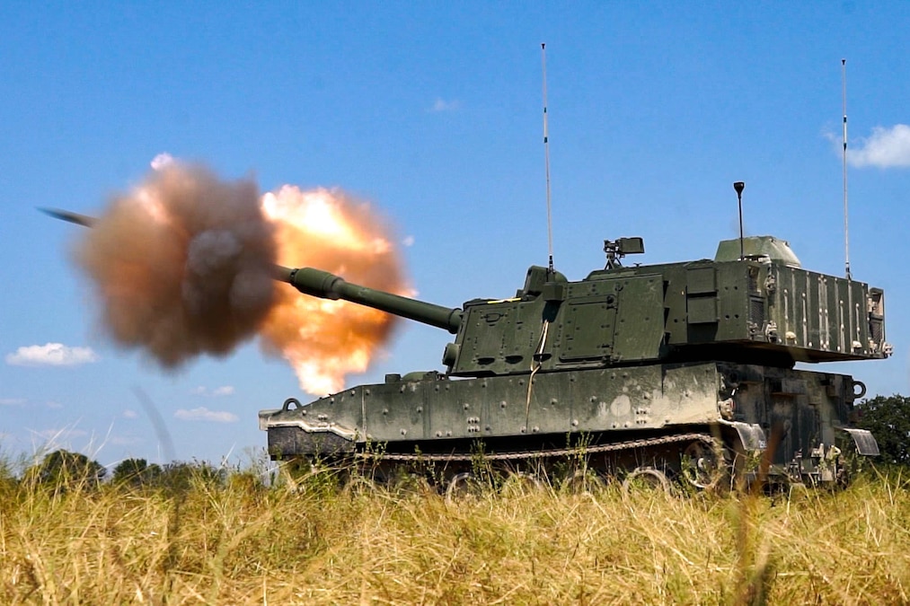Smoke clouds and fire erupt from a green tank as it drives on a yellow field under blue skies.
