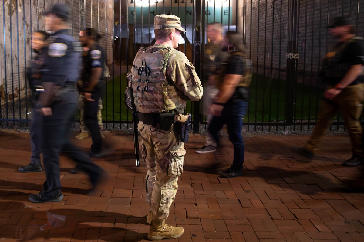 A guardsman stands on brick pavement and watches people wearing law enforcement clothing and gear walk past, with a tall black gate, courtyard and mural painted on a building in the background.