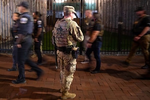 A guardsman stands on brick pavement and watches people wearing law enforcement clothing and gear walk past, with a tall black gate, courtyard and mural painted on a building in the background.