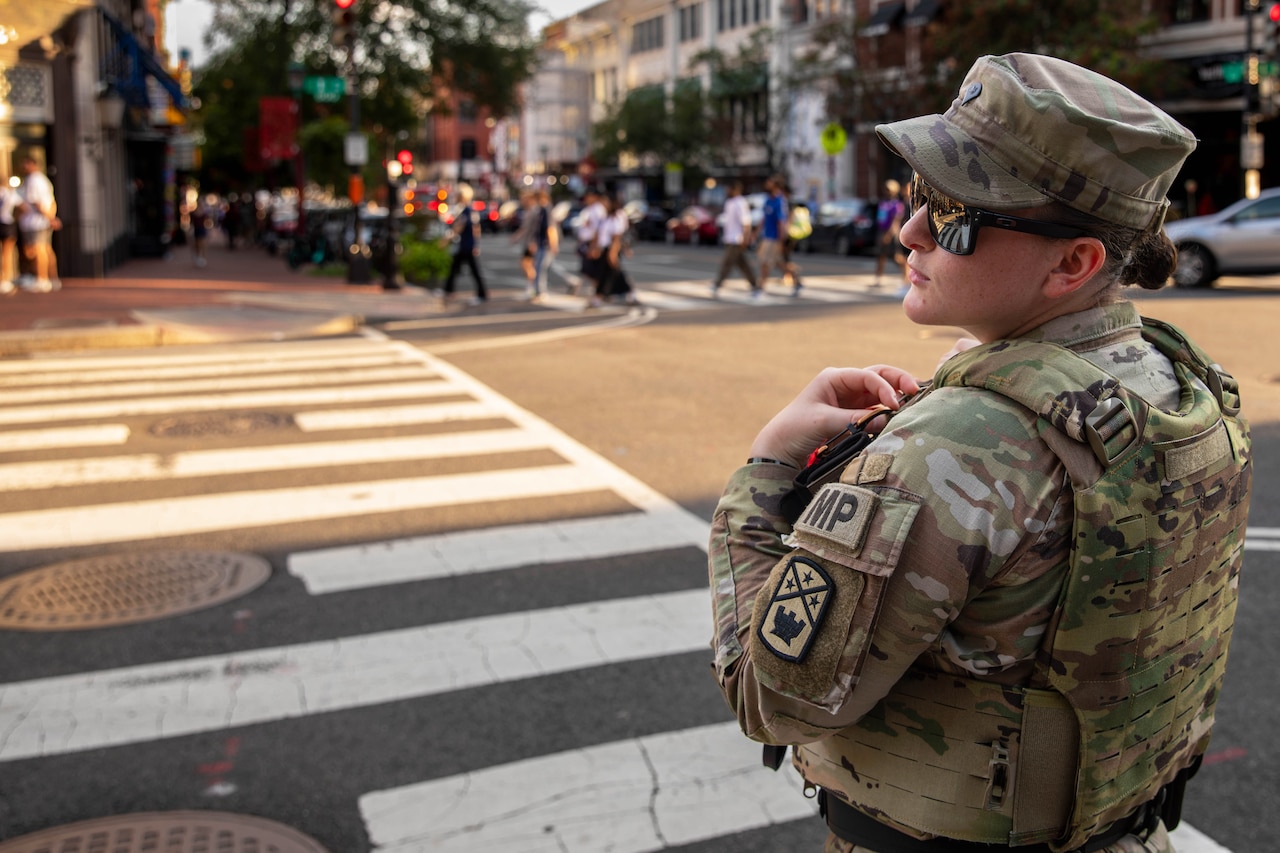 A guardsman stands on a street corner during daytime as pedestrians cross a road lined with buildings and a vehicle waits to turn in the background.