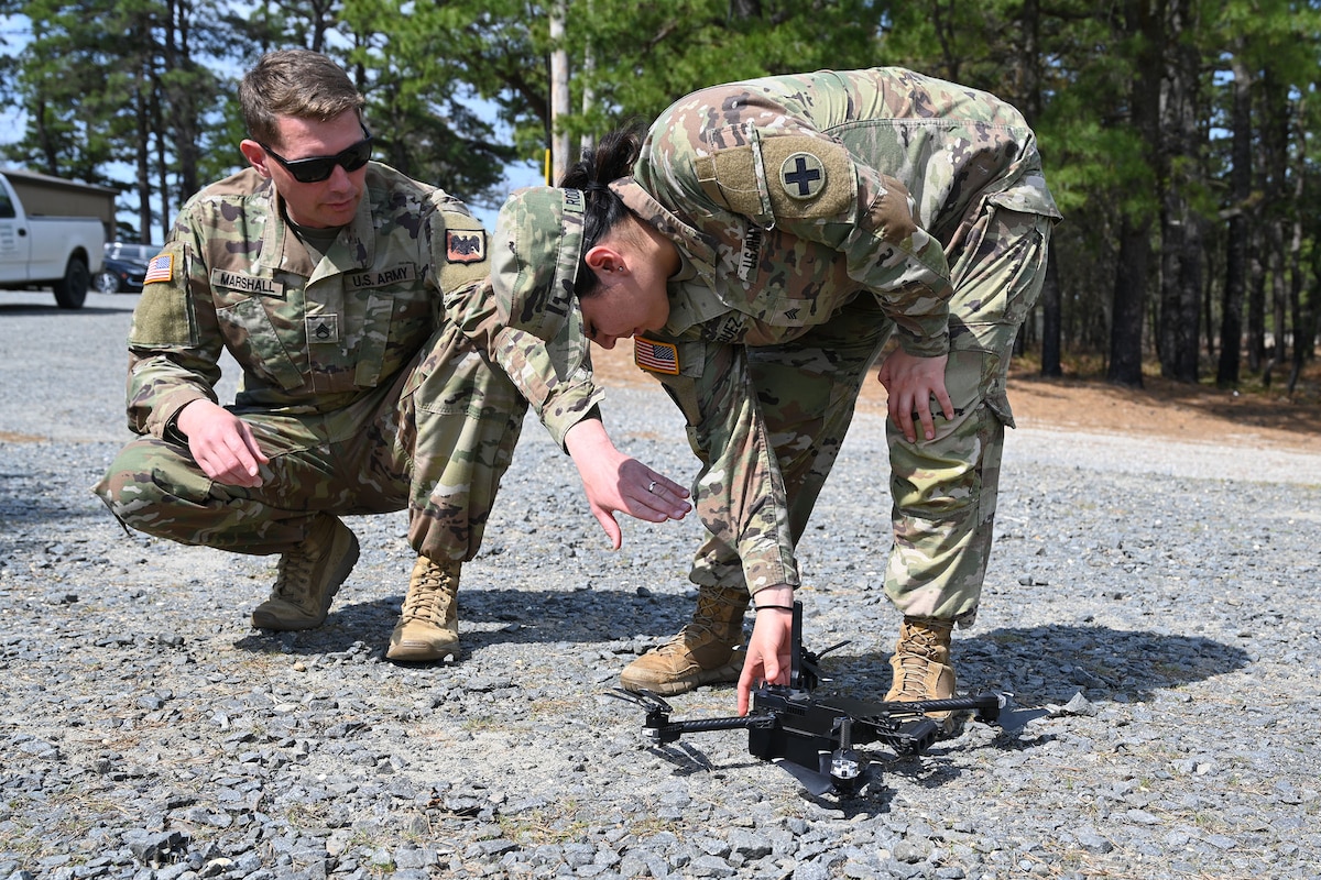 A person wearing a camouflage military uniform stands on a gravel road and attends to a small, unmanned aircraft while another person, wearing similar attire and sunglasses, crouches down and provides assistance.