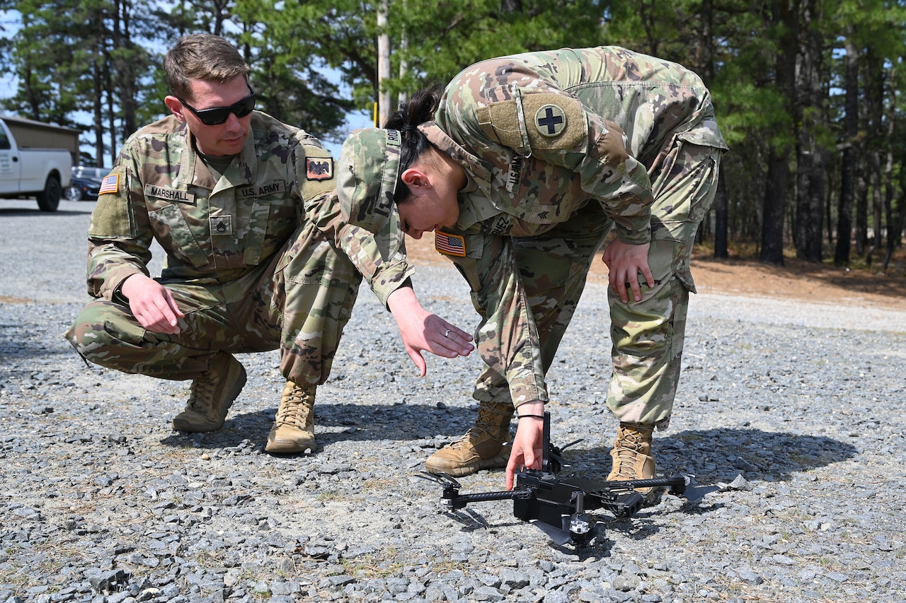 A person wearing a camouflage military uniform stands on a gravel road and attends to a small, unmanned aircraft while another person, wearing similar attire and sunglasses, crouches down and provides assistance.