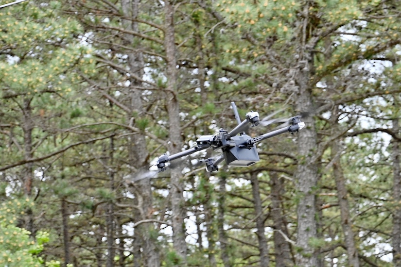 A small, unmanned aircraft flies through the air with a forest in the background.