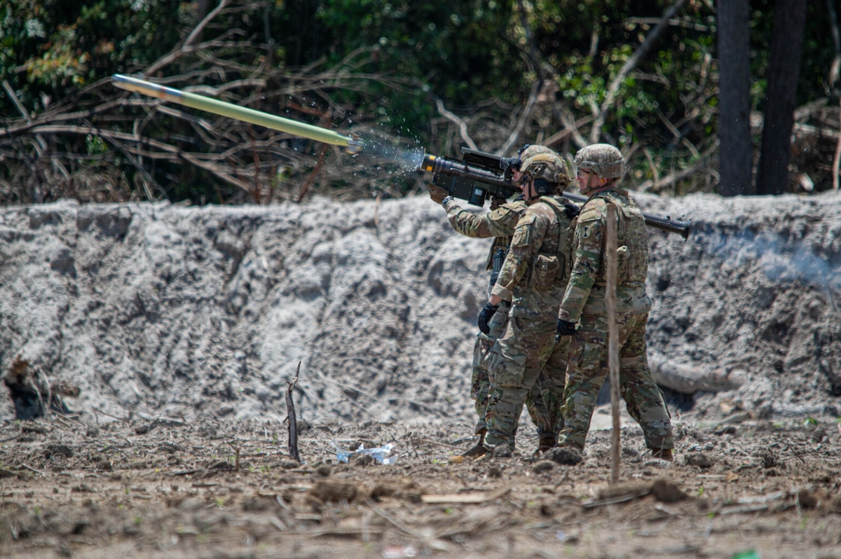 Three men wearing camouflage military uniforms and ballistic helmets stand together in a group as one of the men fires a missile.