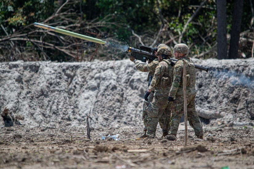 Three men wearing camouflage military uniforms and ballistic helmets stand together in a group as one of the men fires a missile.