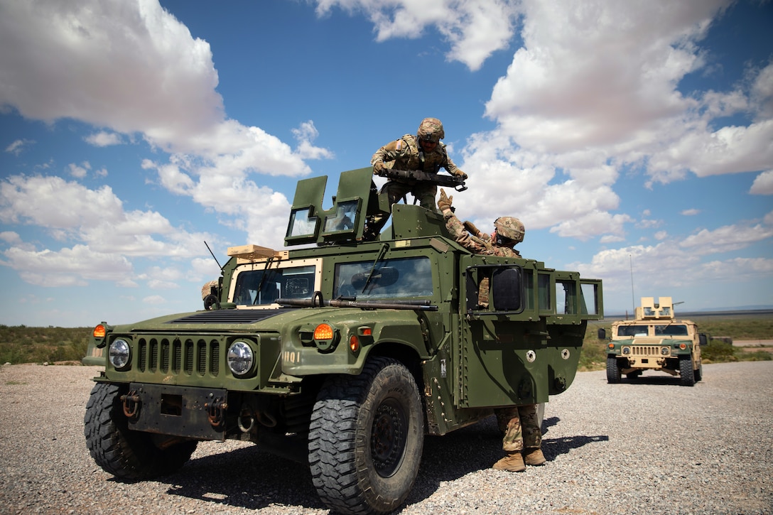 A soldier stands on a Humvee while another soldier hands them a piece of equipment under blue skies dotted with clouds.