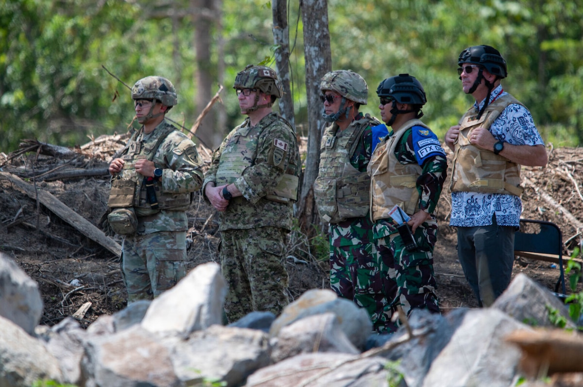 Four men wearing camouflage military uniforms and ballistic helmets, and a man dressed in civilian attire wearing a ballistic helmet, stand together in a group and appear to be observing something that is not in the frame.