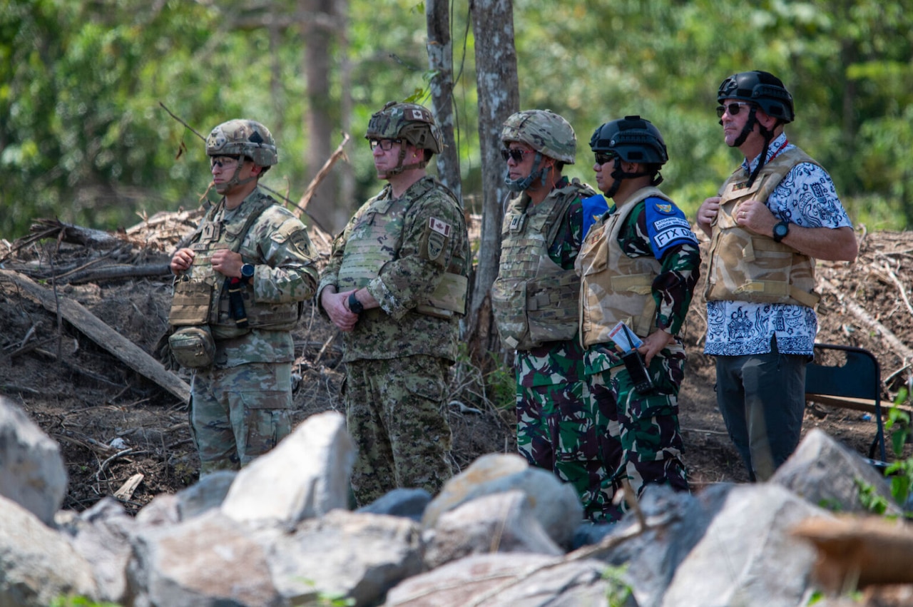 Four men wearing camouflage military uniforms and ballistic helmets, and a man dressed in civilian attire wearing a ballistic helmet, stand together in a group and appear to be observing something that is not in the frame.