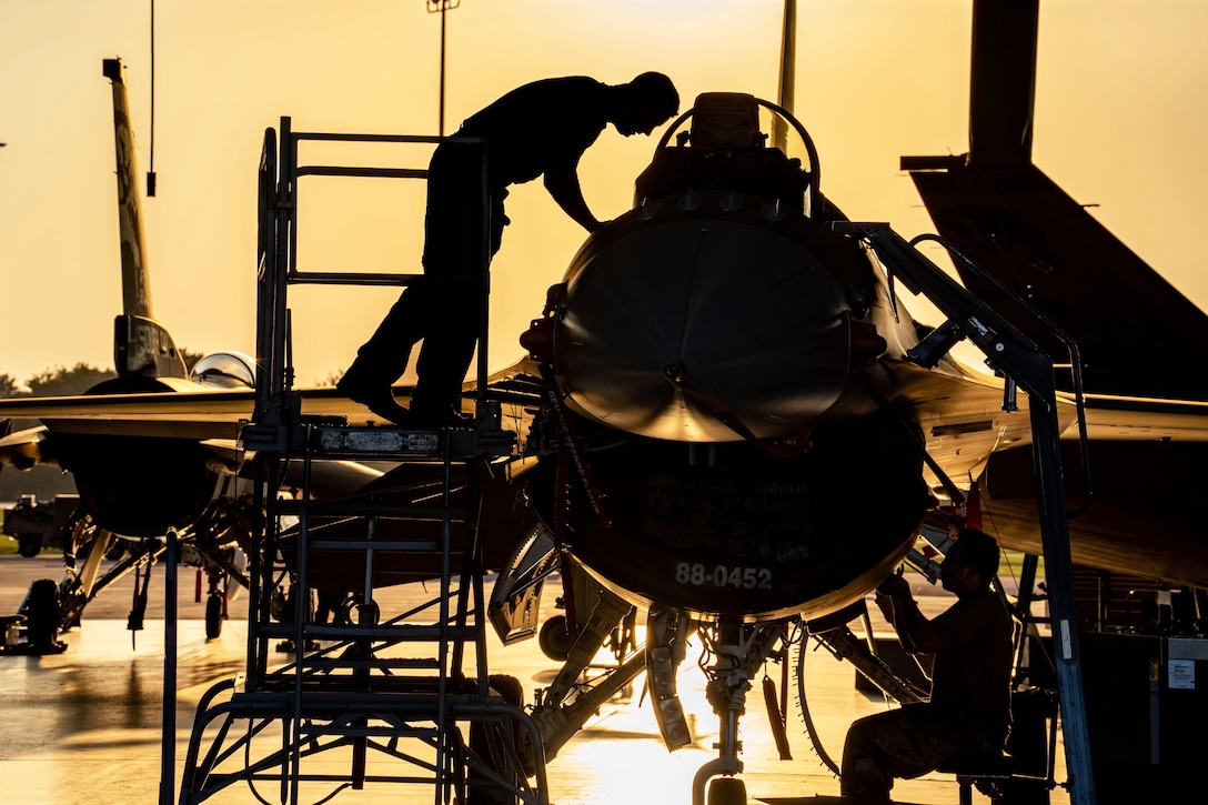 Two airmen in silhouette work on opposite sides of an aircraft in a hangar bay with orangish hues in the background.
