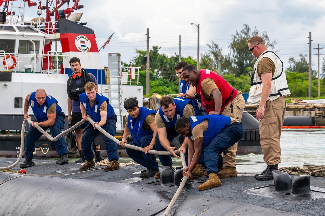 Sailors in blue vests pull a line while standing on top of a submarine with a ship in the background.