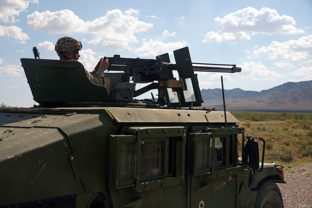 A soldier sits in the cockpit above a Humvee and aims a machine gun in a field under blue skies with clouds.