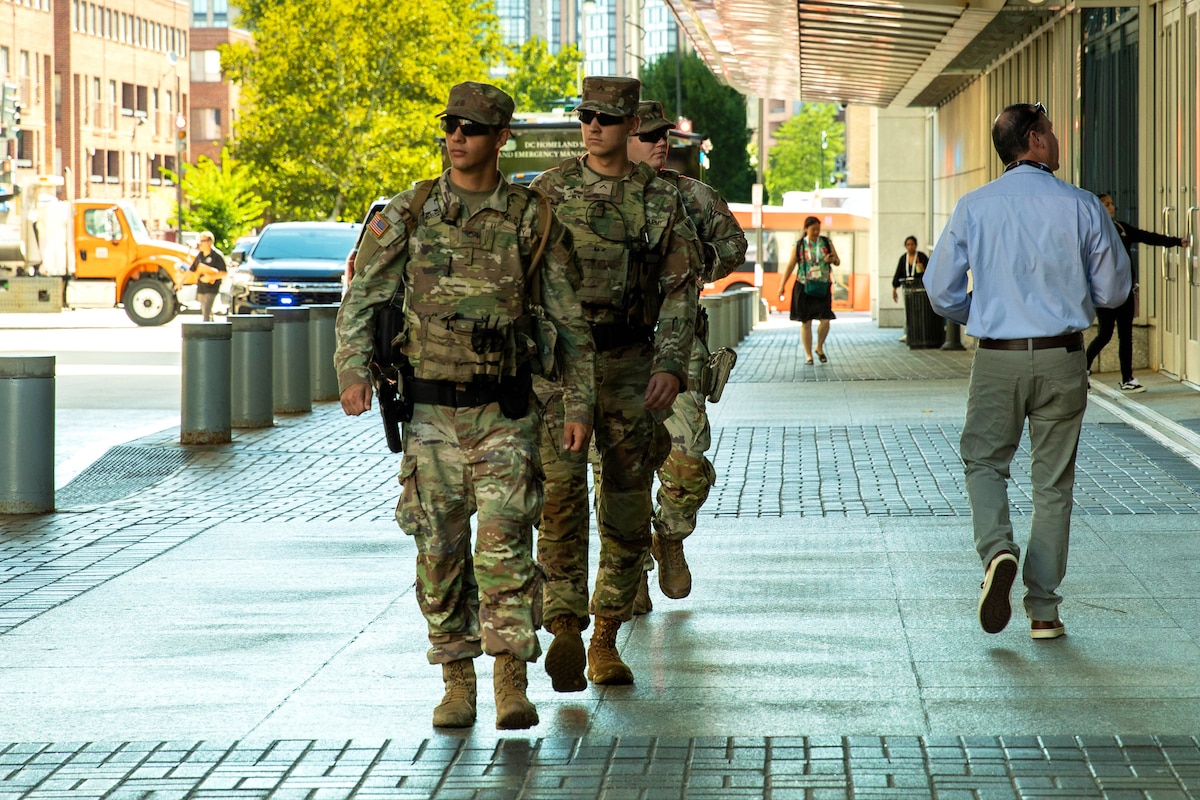 Three guardsmen walk in a line on a sidewalk on a sunny day, with vehicles, buildings, pedestrians and trees in the background.