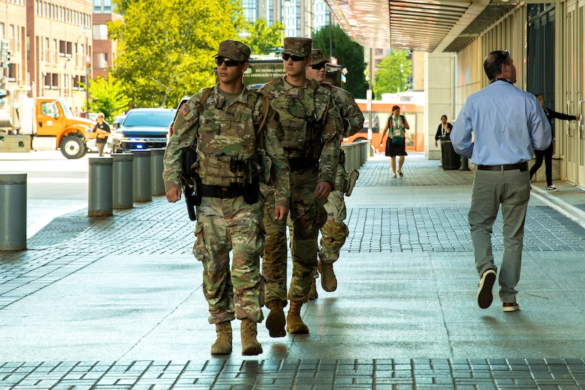 Three guardsmen walk in a line on a sidewalk on a sunny day, with vehicles, buildings, pedestrians and trees in the background.