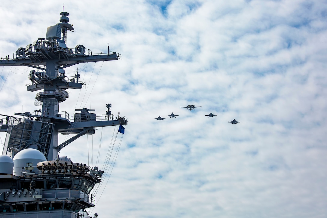 Five aircraft fly in formation toward a cloudy sky as a partially visible ship sits to the left.
