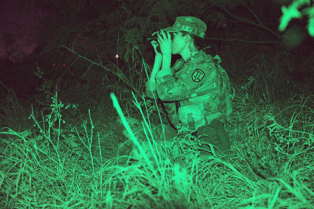 A soldier holds up night vision goggles while standing in tall grass while bathed in green light.