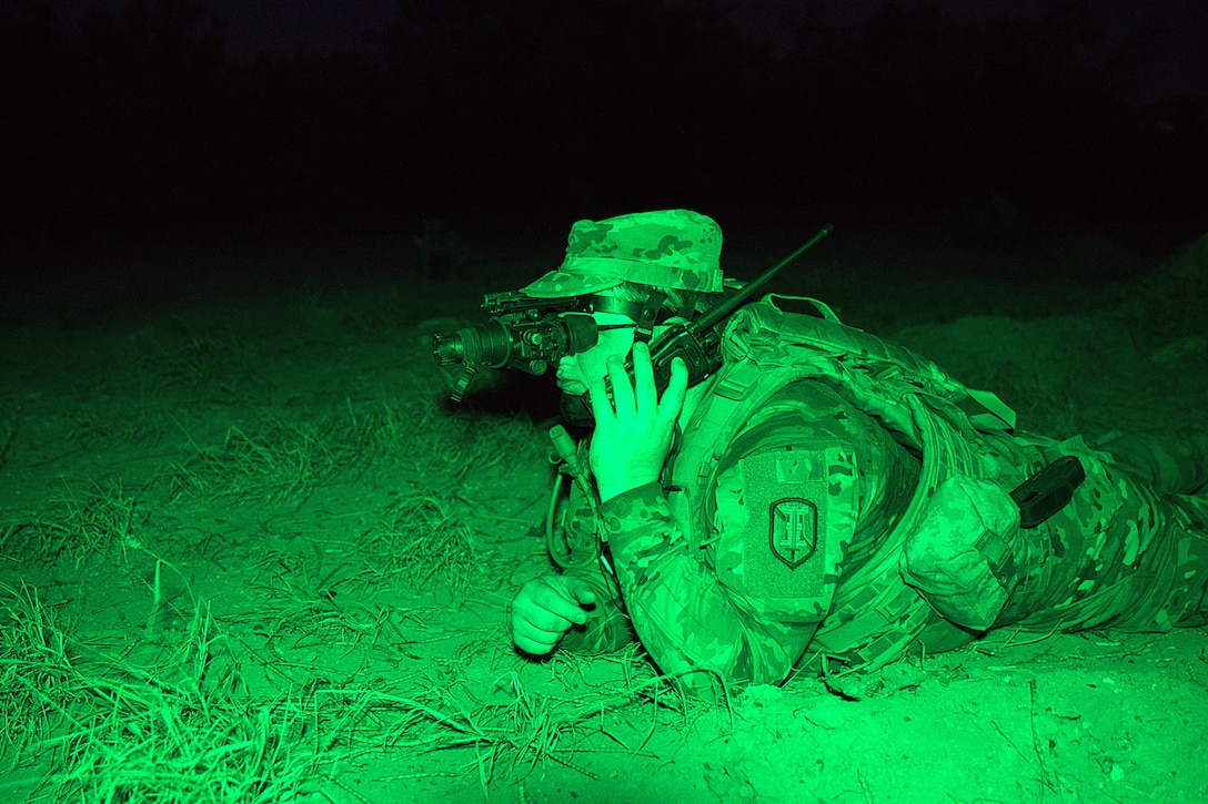 A soldier wearing night vision goggles holds a phone to up to their ear while laying on the ground while bathed in green light.