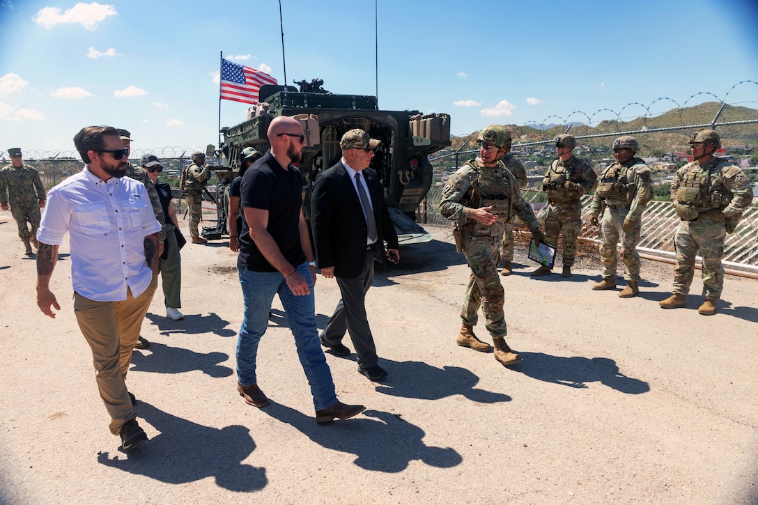 A soldier speaks to three people as they walk by other soldiers on a dirt path outside on a sunny day.