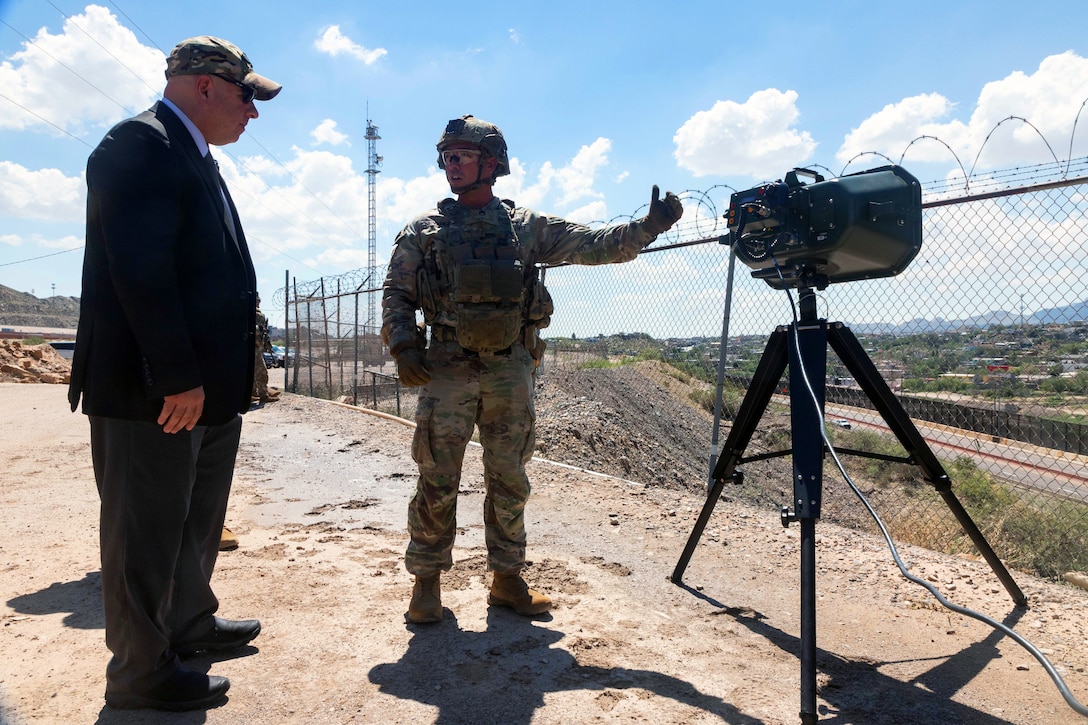 A soldier in tactical gear gestures towards equipment on a tripod while standing next to a person in a suit outside on a sunny day.